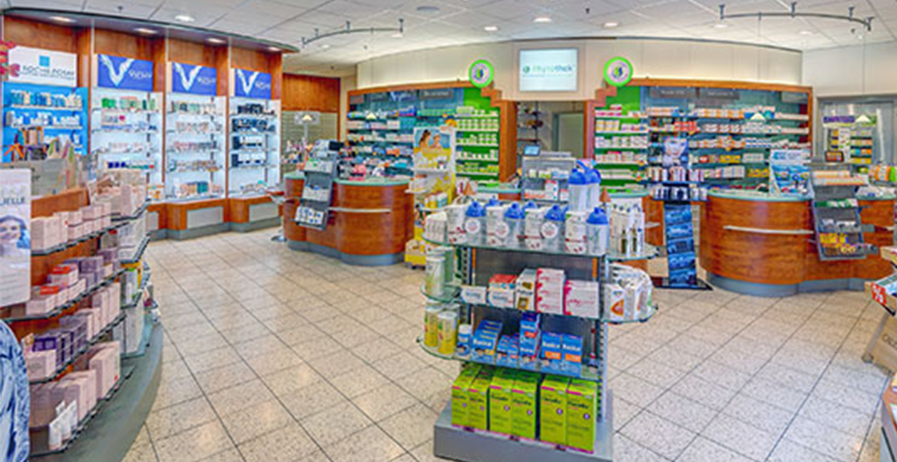 Die Aufnahme zeigt das helle und gut organisierte Innere einer Apotheke. Auf den Regalen sind zahlreiche Medikamente und Gesundheitsprodukte sichtbar. Im Vordergrund stehen mehrere Theken und Regale mit Produktdisplay, und an den Wänden sind weitere Regale mit Medikamenten zu sehen. Die Beleuchtung ist hell und gleichmäßig, was die Produkte gut sichtbar macht. Der Raum wirkt modern und einladend.
