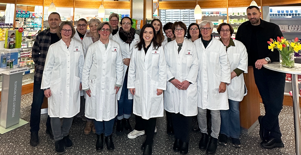Gruppenfoto des Teams einer Apotheke. Mehrere Frauen in weißen Apothekenkitteln stehen zusammen und lächeln in die Kamera. Zwei Männer in dunkler Kleidung stehen jeweils links und rechts am Bildrand. Im Hintergrund sind Regale mit Arzneimitteln und Produkten sowie ein Aufsteller mit der Aufschrift „Produkt des Monats“ zu sehen. Auf einem Tisch rechts im Bild steht ein Strauß mit gelben und roten Tulpen.