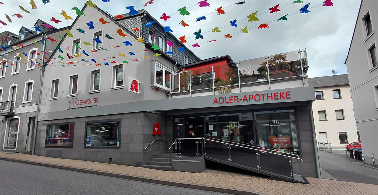 Außenansicht der Adler-Apotheke von der Straßenseite. Das Gebäude ist modern gestaltet mit grauer Fassade, großen Schaufenstern und einem gut sichtbaren Apotheken-Logo. Über dem Eingang befindet sich eine rote Beschriftung „ADLER-APOTHEKE“. Eine Rampe und Treppe mit Handläufen führen barrierefrei zum Eingang. Über der Straße hängen bunte Dekofahnen in Tierform.