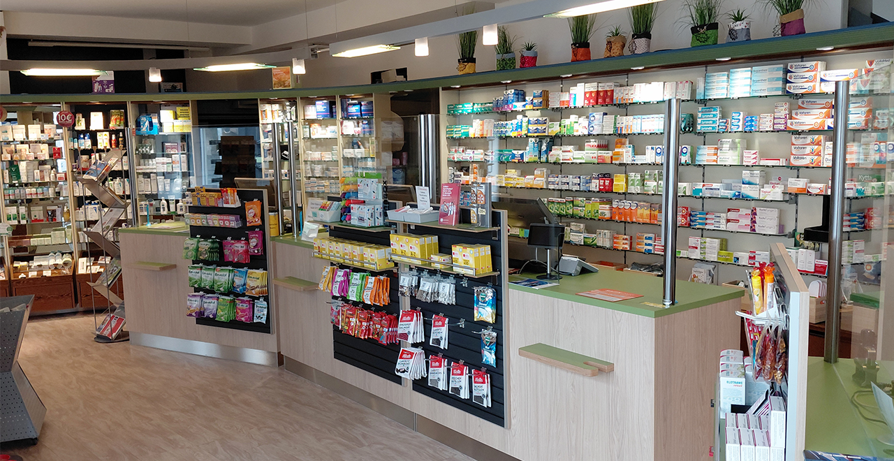Innenansicht der Adler-Apotheke mit modernem Verkaufsbereich. Mehrere bedienbare Theken mit Plexiglasschutz, gut sortierte Regale mit Medikamenten und Produkten, sowie dekorative Pflanzen auf der oberen Regalfläche. Freundliche und helle Atmosphäre mit klarer Struktur.
