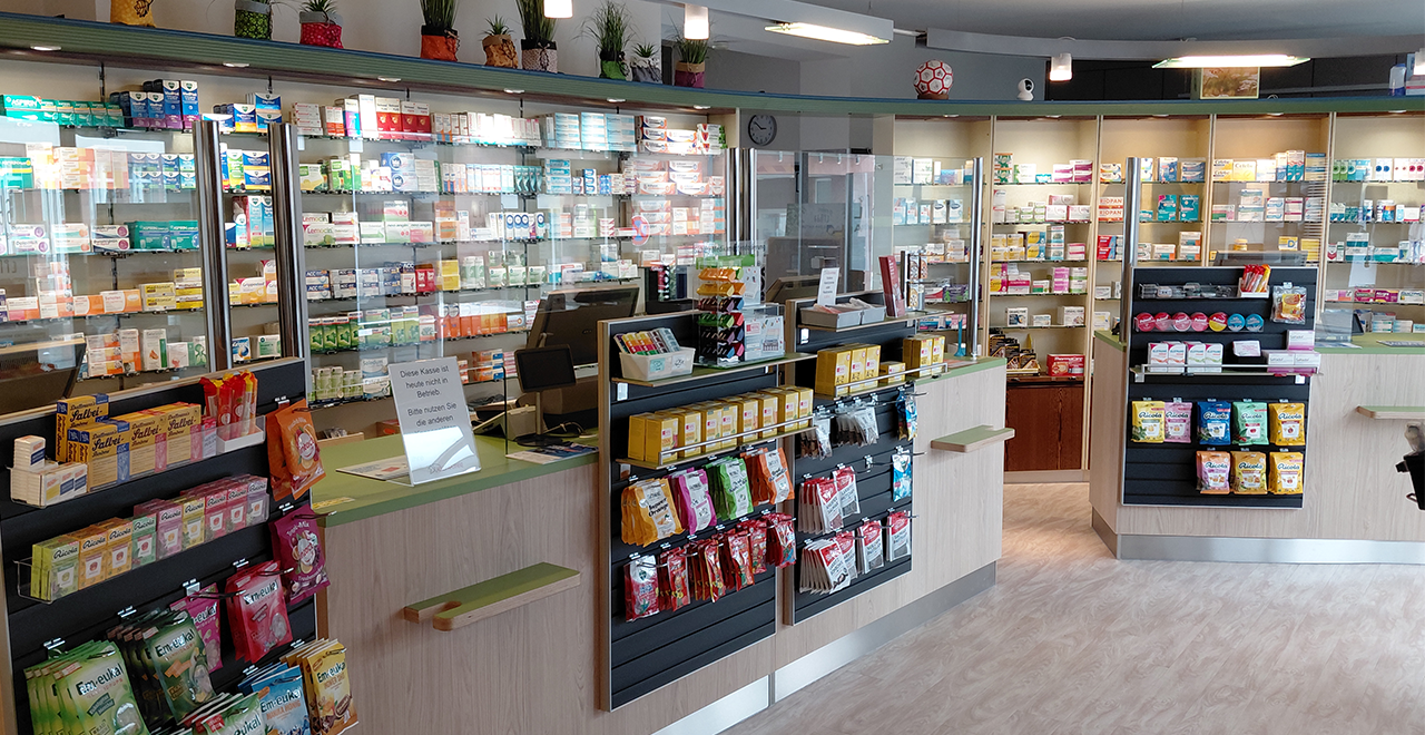 Blick in eine helle, moderne Apotheke mit Holzregalen und Verkaufstheken. Die Theken sind mit einer gläsernen Trennwand ausgestattet. Auf den Regalen und hinter der Theke stehen zahlreiche Arzneimittel und Pflegeprodukte. Der Boden ist mit hellem Holzfußboden ausgelegt. Kleine Pflanzen stehen als Dekoration auf den oberen Regalen. Die Apotheke wirkt ordentlich, sauber und einladend.