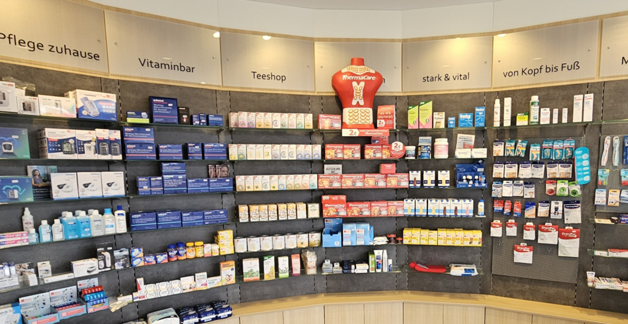Innenaufnahme der Apotheke mit Blick vom Eingang aus. Links befinden sich Wartebereich und Süßwarenständer, geradeaus mehrere Verkaufstheken mit Plexiglas und Produktregalen.