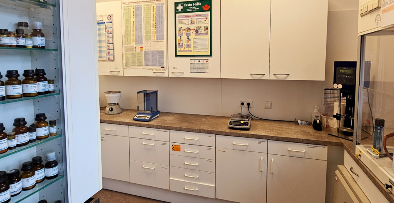 Apothekenlabor mit weißen Schränken, Laborwaagen, Chemikalien und einem Erste-Hilfe-Poster an der Wand. Auf der linken Seite ein Regal mit braunen Reagenzgläsern.