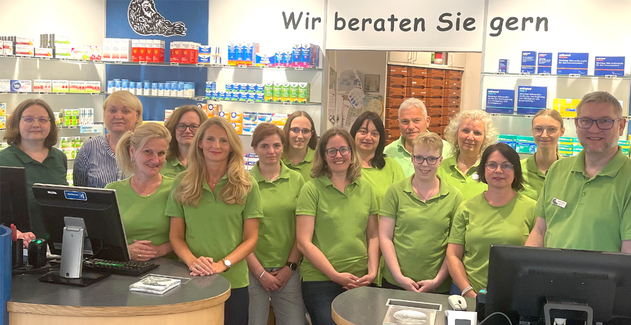 Gruppenfoto eines Apotheken-Teams mit 16 Personen, die überwiegend grüne Polo-Shirts tragen. Sie stehen hinter dem Verkaufstresen, umgeben von Regalen mit Medikamenten. Über ihnen hängt ein Schild mit der Aufschrift: „Wir beraten Sie gern“.