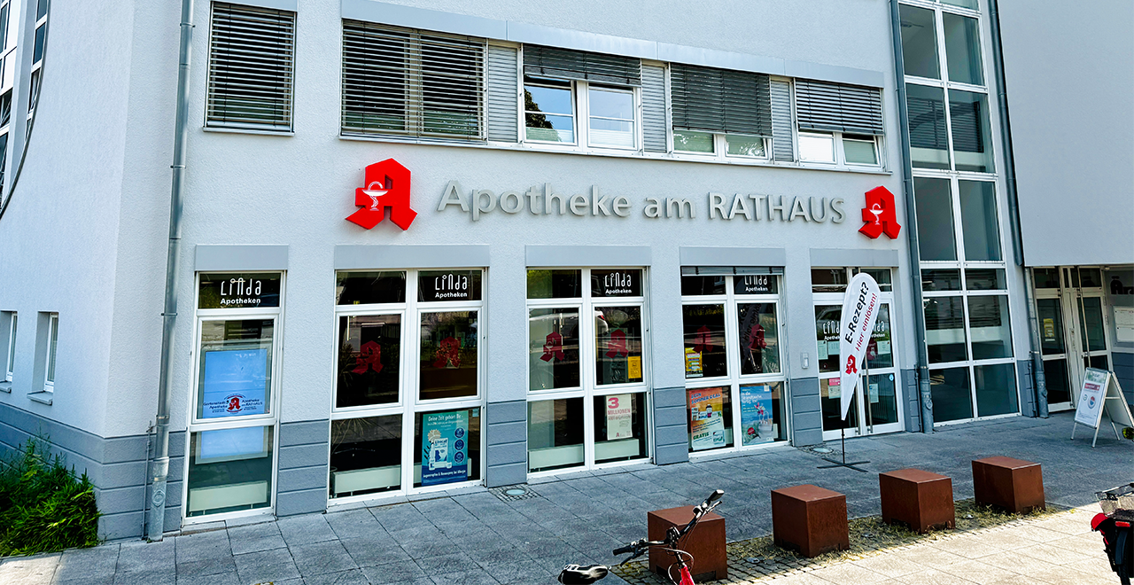 Moderne Außenansicht der „Apotheke am RATHAUS“ in einem hellen Gebäudekomplex. Über der Glasfassade steht der Apothekenname in silbernen Buchstaben, daneben befindet sich das rote Apotheken-A mit Schale und Schlange. Mehrere Fenster zeigen das „Linda Apotheken“-Logo. Eine weiße Beachflag mit der Aufschrift „E-Rezept? Wir sind bereit!“ steht vor dem Eingang. Ein Fahrrad und Sitzwürfel befinden sich auf dem Gehweg.