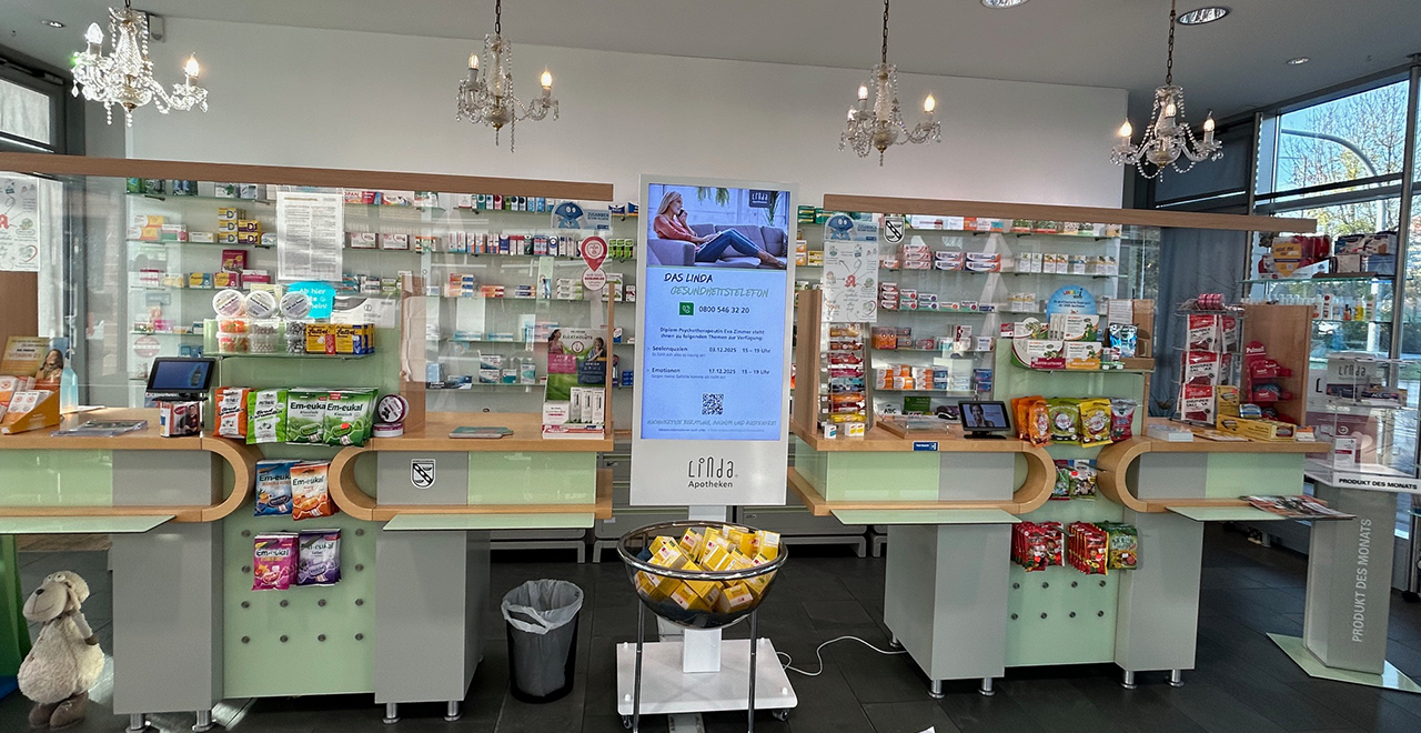Innenraum einer Apotheke mit drei Bedienplätzen, Regalen voller Arzneimittel und einem digitalen Bildschirm in der Mitte.