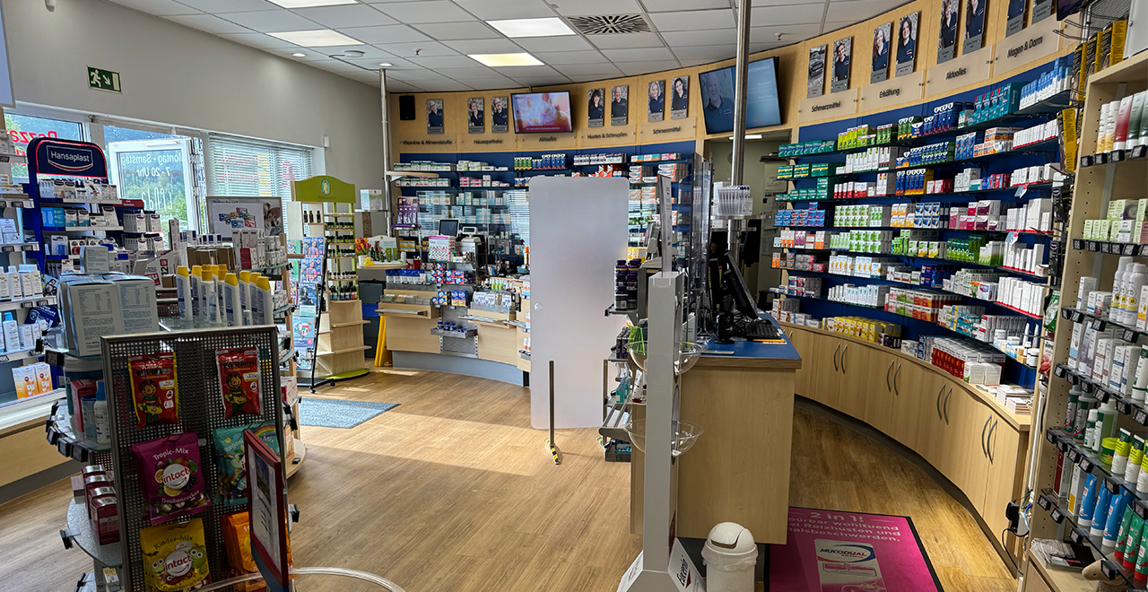 Heller, moderner Verkaufsraum der Pazzawa-Apotheke mit Regalen voller Medikamente und Pflegeprodukte, sowie Porträts des Teams an der Wand.