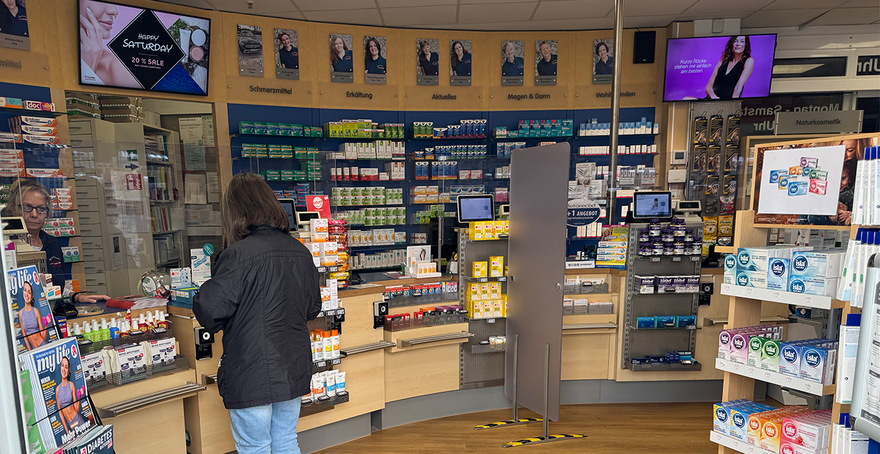 Kundin im Gespräch mit einer Mitarbeiterin an der Apothekenkasse, umgeben von Medikamentenregalen und Werbebildschirmen.