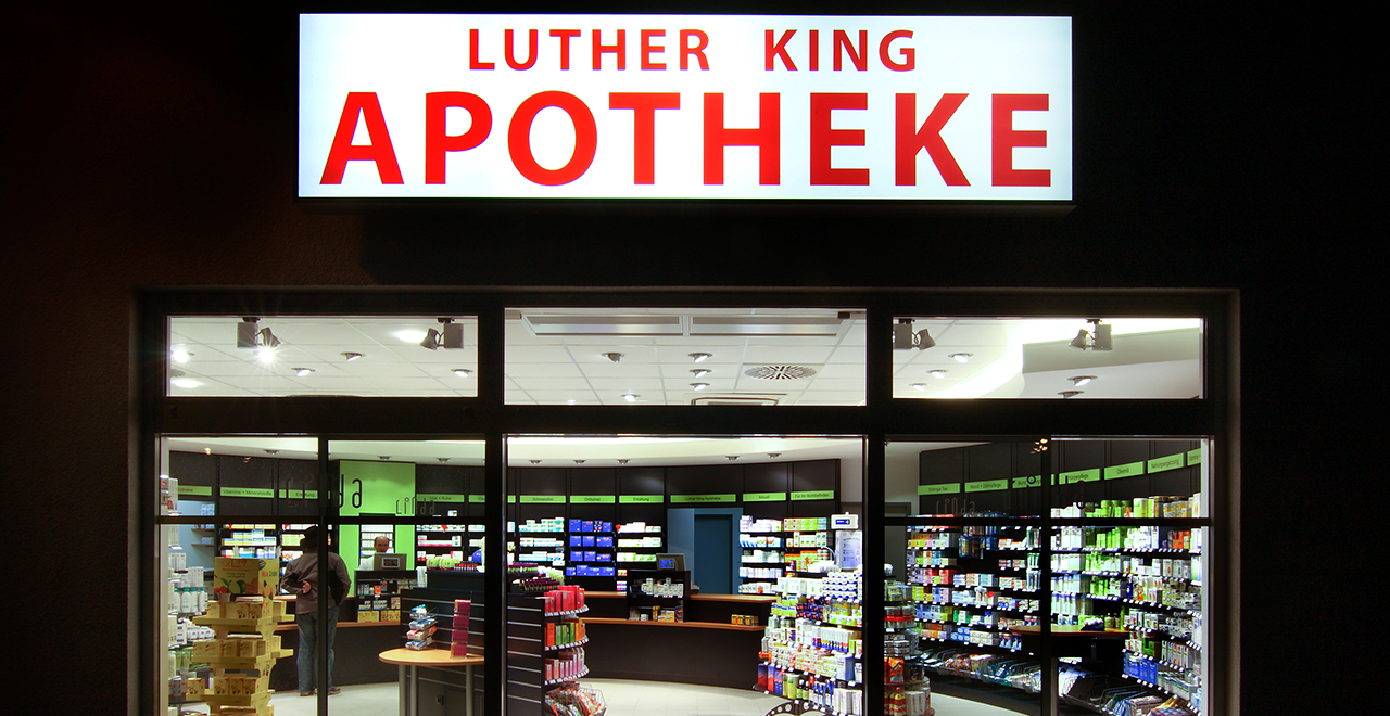 Nachtaufnahme der „Luther King Apotheke“ mit großem, beleuchtetem Schild über dem Eingang. Durch die großen Glasfenster ist der helle Innenraum mit Regalen und Kunden sichtbar.