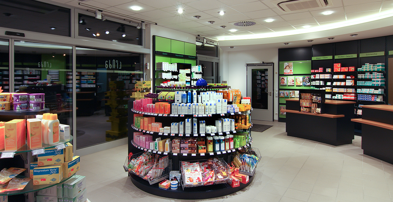 Innenansicht einer modernen Apotheke mit hell ausgeleuchtetem Verkaufsraum. In der Mitte steht ein rundes Regal mit Hautpflegeprodukten, Nahrungsergänzungsmitteln und Kosmetika. Im Hintergrund sind weitere Regale mit Arzneimitteln und Produkten des täglichen Bedarfs zu sehen.
