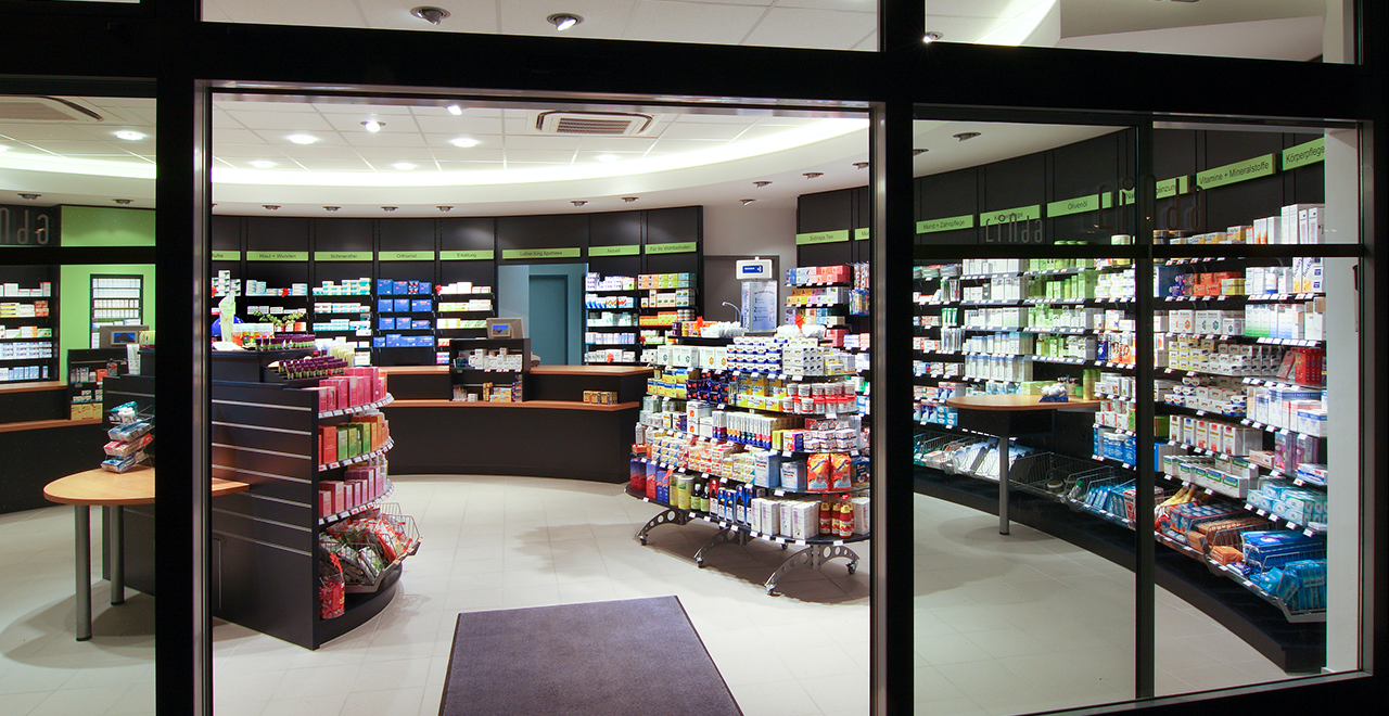 Blick durch das Schaufenster in den Innenraum einer Apotheke bei Nacht. Die Verkaufsfläche ist gut beleuchtet und zeigt eine Vielzahl an Produkten, darunter Medikamente, Pflegeprodukte und Gesundheitsartikel. Die Regale sind ordentlich und übersichtlich angeordnet.