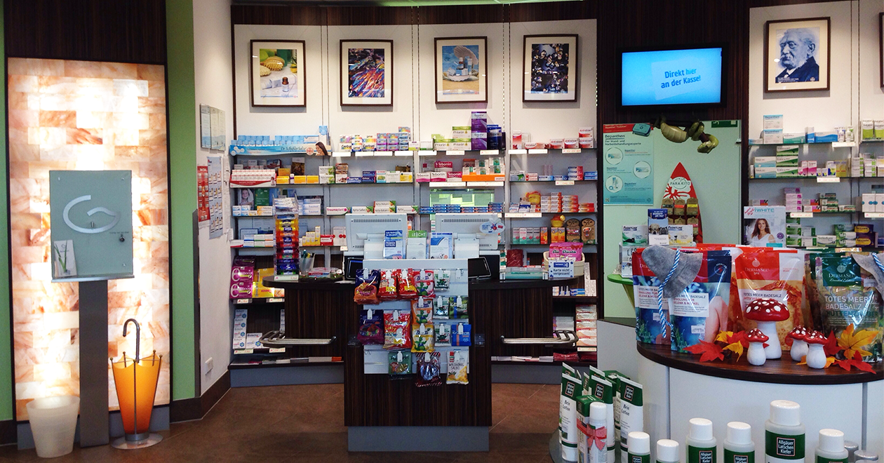 Innenansicht einer Apotheke mit einem gut sortierten Verkaufsbereich. Im Mittelpunkt stehen zwei Kassentresen mit Produkten und Schutzscheiben. Im Hintergrund befinden sich Regale mit Medikamenten, dekorative Bilder an der Wand und ein Bildschirm mit der Aufschrift „Direkt hier an der Kasse!