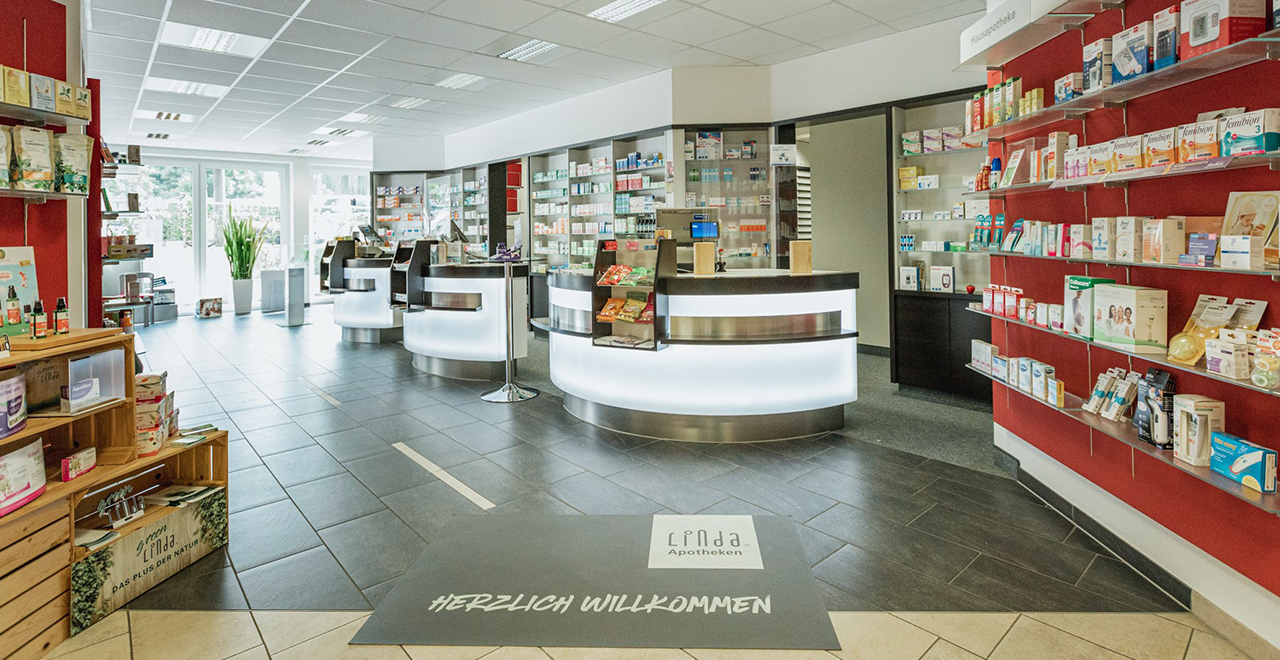 Heller Innenraum einer Apotheke mit modernen, halbkreisförmigen Verkaufstresen und zahlreichen Medikamentenregalen. Auf dem Boden liegt eine Fußmatte mit der Aufschrift 