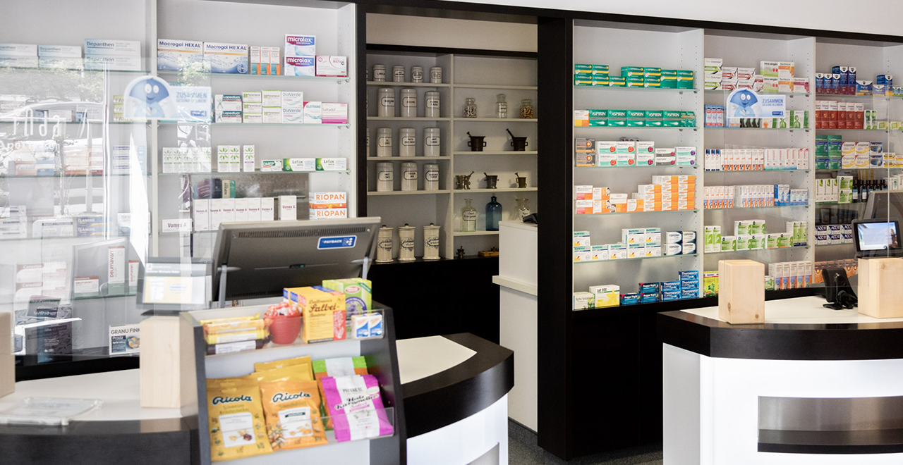 Blick auf den Verkaufsbereich einer Apotheke mit drei modernen, beleuchteten Tresen, vielen Arzneimitteln in Wandregalen und einer auffälligen roten Präsentationswand mit Produkten.