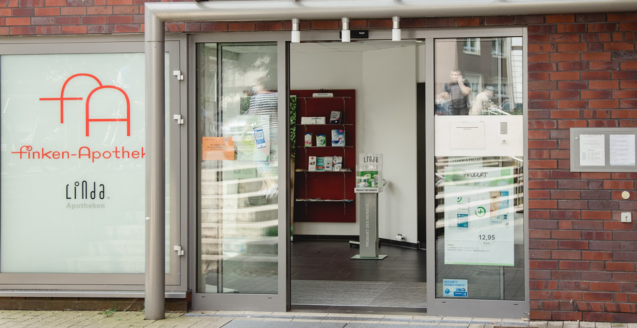 Nahaufnahme der Apothekentheke mit Sicht auf die Kasse und eine Auswahl an rezeptfreien Produkten wie Ricola Bonbons. Im Hintergrund sind Arzneimittelregale und traditionelle Apothekengefäße sichtbar.