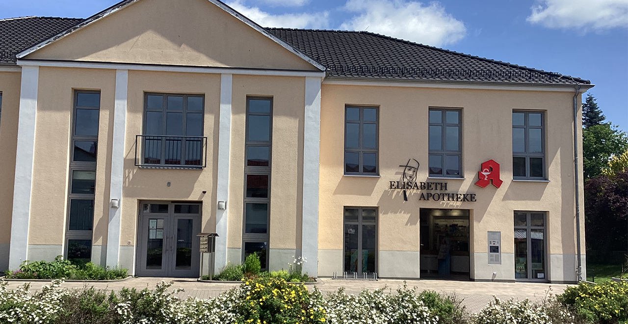 Frontalansicht des Gebäudes mit dem Eingang der „Elisabeth Apotheke“. Über der Tür befindet sich der Schriftzug sowie ein rotes Apotheken-Symbol. Vor dem Gebäude sind Pflanzen und Blumen angelegt, und die Fassade ist in hellen Beige- und Grautönen gehalten.