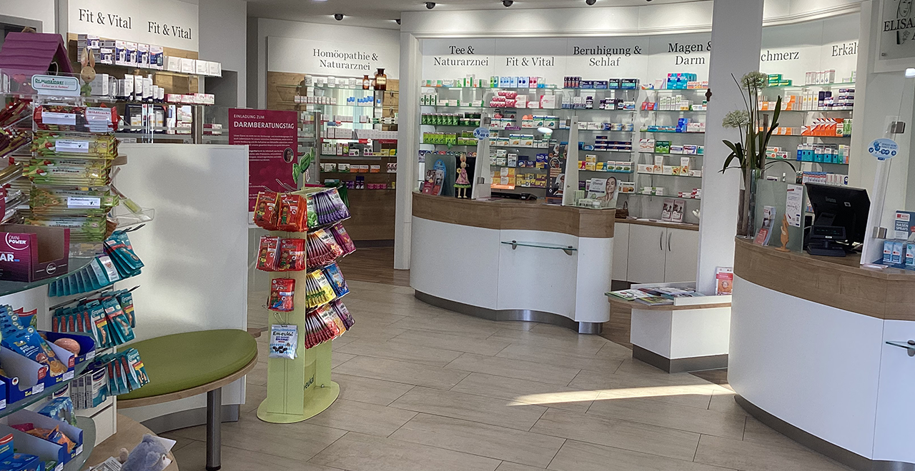 Heller, moderner Verkaufsraum einer Apotheke. Im Vordergrund stehen Regale mit verschiedenen Produkten, im Hintergrund ein geschwungener Verkaufstresen mit Plexiglasschutz. An den Wänden sind übersichtlich angeordnete Medikamente und Gesundheitsprodukte nach Kategorien wie „Fit & Vital“, „Beruhigung & Schlaf“ und „Magen & Darm“.