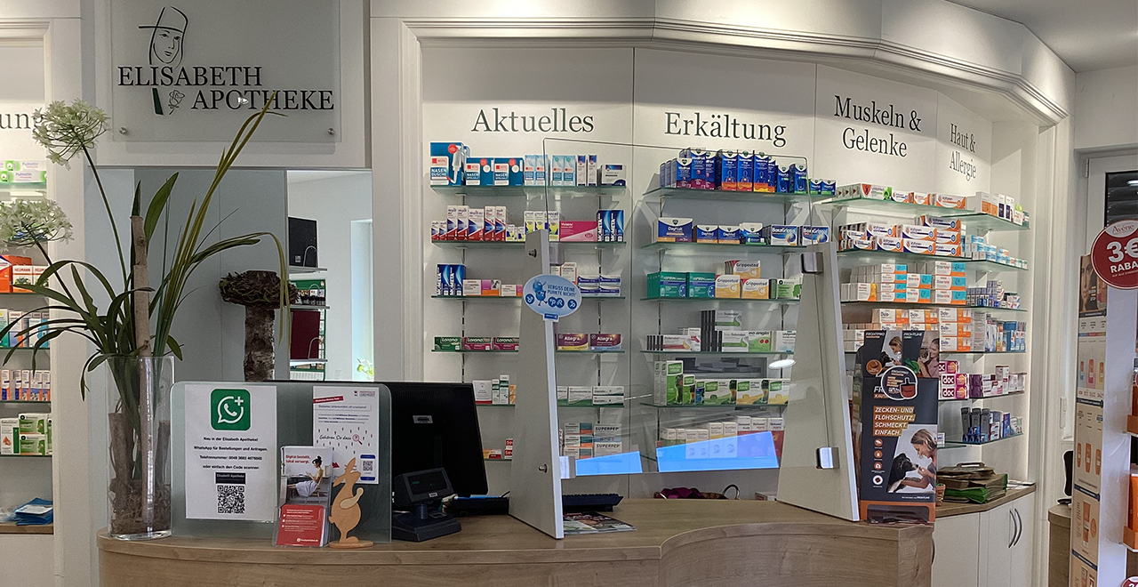 Detailansicht des Kassen- und Beratungsbereichs einer Apotheke. Hinter dem Tresen befinden sich Regale mit Medikamenten, beschriftet mit Kategorien wie „Aktuelles“, „Erkältung“, „Muskeln & Gelenke“ sowie „Haut & Allergie“. Auf dem Tresen stehen ein Computer, Informationsmaterial und eine Pflanze.