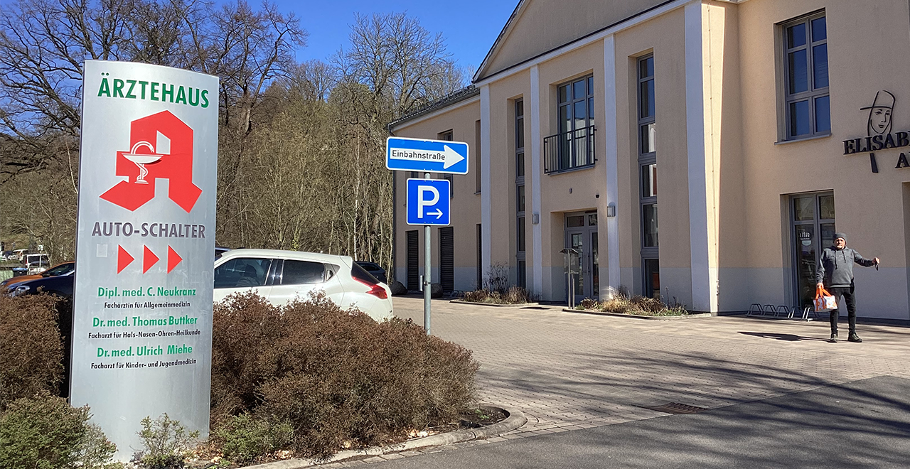 Außenansicht eines modernen, zweigeschossigen Gebäudes mit heller Fassade und großen Fenstern, in dem sich eine Apotheke befindet. Links im Vordergrund steht ein Schild mit der Aufschrift „Ärztehaus“ und einem roten Apotheken-Symbol sowie dem Hinweis „Auto-Schalter“ , daneben sind Parkplätze.