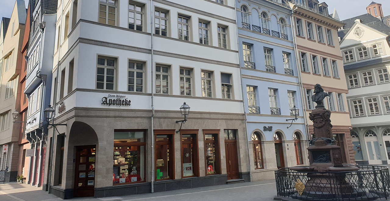 Straßenszene in einem historischen Stadtviertel mit renovierten Häuserfassaden in verschiedenen Farben. Im Vordergrund eine Apotheke mit dem Schriftzug „Dom Römer Apotheke“ im Erdgeschoss eines weißen Gebäudes. Rechts davon ein kleiner Platz mit einem Brunnen und einer Bronzebüste. Die Architektur zeigt klassische europäische Stile mit vielen Fenstern, Erkern und dekorativen Details. Keine Menschen sichtbar, die Straße wirkt ruhig und sauber.